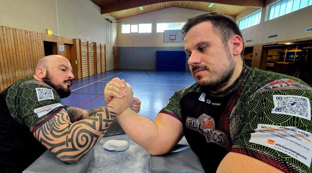Zwei Armwrestler der Pulldogs Germany am Tisch, im Hintergrund die leere Sporthalle